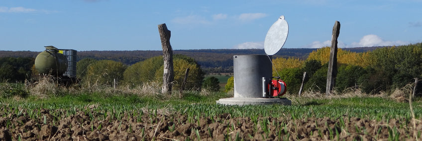 At the edge of a field, a gray cylinder with an open lid stands out of the ground.